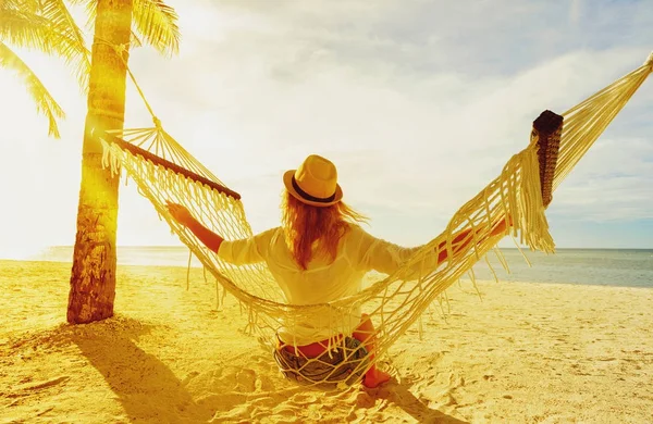 Woman in hat  sitting on hammock between palms on the beach and enjoying sunset.