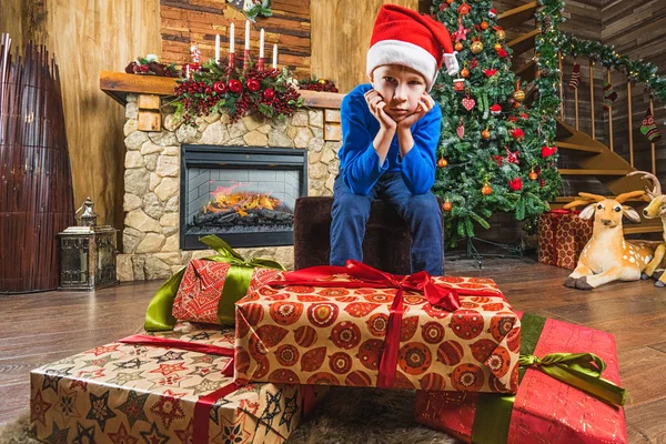 Very sad child with christmas gift boxes. Fireplace and Christmas tree on background. Merry Christmas and New Year concept