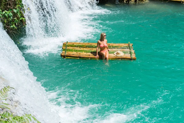 Woman in bikini sitting on bamboo raft close to waterfall.