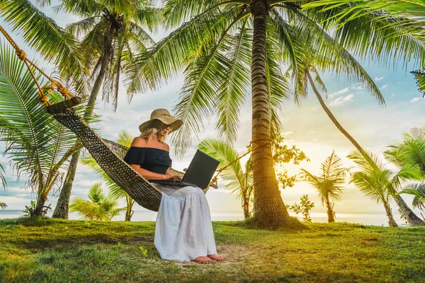 妇女在帽子坐在吊床在海滩与膝上型电脑 — 图库照片