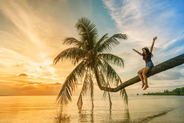 妇女在帽子坐在海滩手掌与举起的手和看美丽的日落 — 图库照片