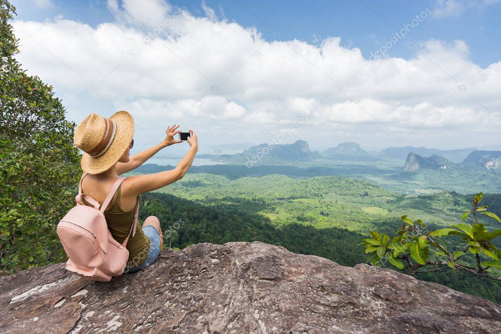 Tourist taking picture on her smartphone from top of mountain. Woamn in hat and with backpack sitting on rock. Travel concept.