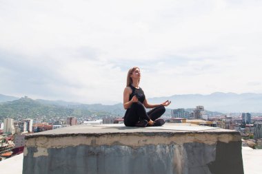 Çatıdaki minderde yoga yapan bir kadın. Yoga ve meditasyon yapan bir kız. Arka planda şehir ve dağlar.
