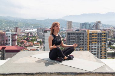 Çatıdaki minderde yoga yapan, yoga ve meditasyon yapan bir kadın. Arka planda şehir ve dağlar.