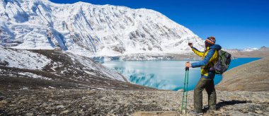 Seyahat ve zeka konsepti: sırt çantalı ve akıllı telefonlu Trekker Tilicho gölünde. Dağların karlı zirveleri ve arka planda Tilicho Gölü. Pankart sürümü.