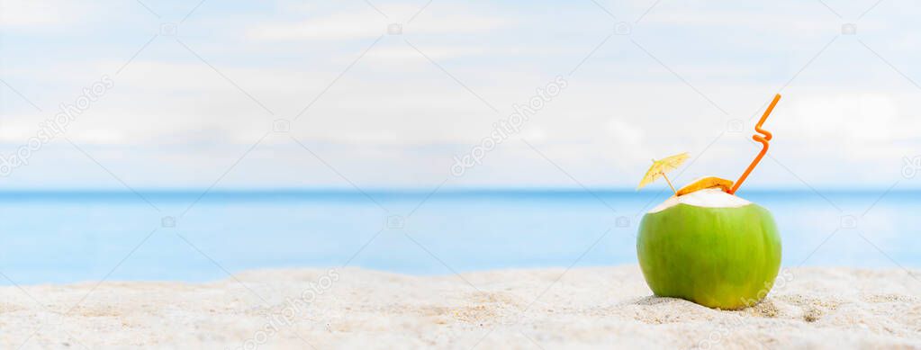 Fresh young coconut lying on the sand beach and blue sky background with straw and umbrella, ready for drink. Tropical vacation travel concept. Copy space.  Focus on coconut. Banner edition.