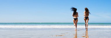 Two young women in swimwear running on the beach. Back view. Hair in motion view. Banner edition.	