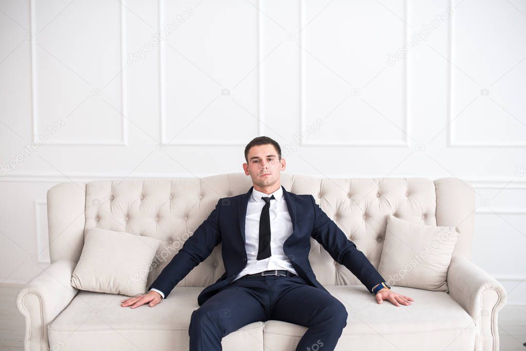 Portrait of a young serious and confident man in a suit sitting on the couch and looking at camera.