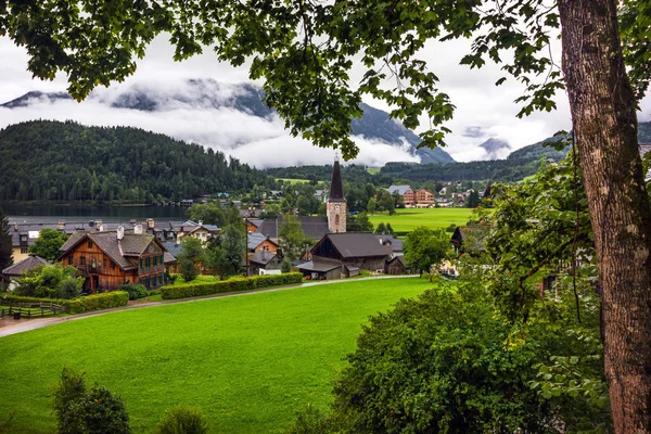 Zielone pastwiska z alpejskiej wioski Altaussee przez jezioro Altausseersee w deszczowy poranek z chmury i mgliste góry w tle — Zdjęcie stockowe