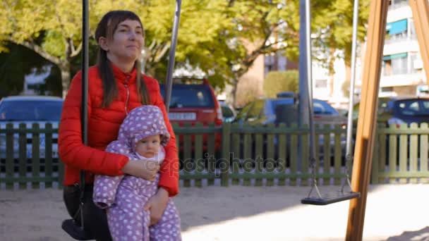 Mãe segurando o bebê em seus braços balançando no balanço no parque de outono — Vídeo de Stock