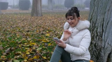 Gülümseyerek, sonbahar Park düşen sarı yapraklar üzerinde oturan Galeri akıllı telefon, tarama güzel kadın.