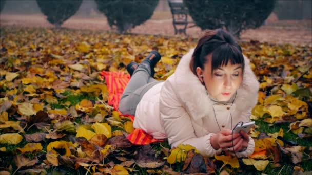 若い女性が秋の公園で黄色と赤の落ち葉の上に横たわる電話で命令を与える. — ストック動画