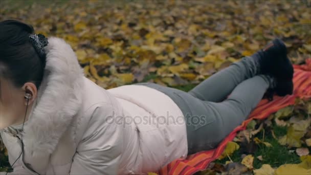 Young woman passionately talking on the phone with headphones lying on fallen leaves in autumn park. — Stock Video