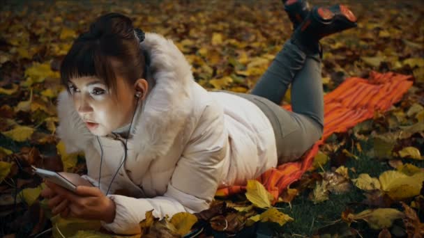 ひたすら秋の公園で落ち葉の上に横たわるヘッドフォンを通して電話で話している若い女性. — ストック動画