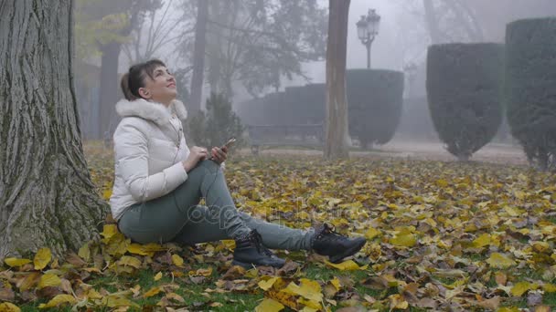 Натхненна дівчина дивиться вгору, посміхається, тримає смартфон і сидить на опале листя в осінньому парку — стокове відео