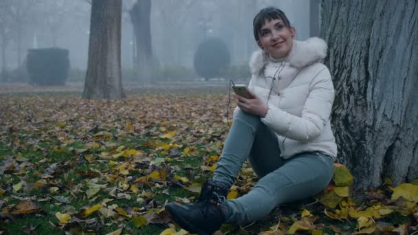 Bella donna pensando, sorridente, seduto a terra sotto l'albero nel parco nebbioso autunno . — Video Stock