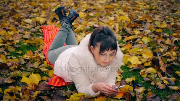 かわいい女の子を驚かせた秋の公園で黄色の落ち葉の上に横たわるスマート フォンの画面を見て. — ストック動画