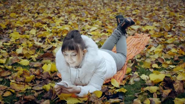 Chica bonita sorprendida mirando la pantalla del teléfono inteligente acostada sobre hojas amarillas caídas en el parque de otoño . — Vídeo de stock