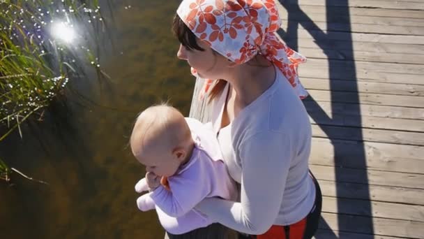 Мама держит ребенка сидя на перилах моста через озеро — стоковое видео