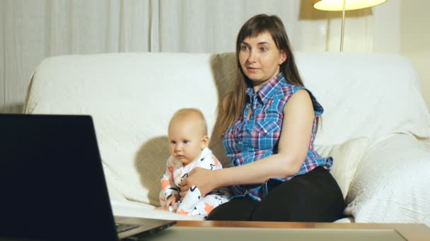 Madre y bebé de ojos azules sentado en un sofá blanco viendo video en la pantalla del ordenador portátil — Vídeos de Stock