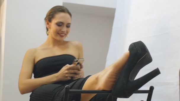 Ontspannen zakenvrouw in zwarte avondjurk zit een hoge hak schoen met behulp van smartphone en glimlachend bungelen — Stockvideo