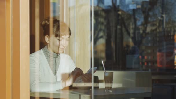 Νεαρή γυναίκα πληκτρολογώντας ένα μήνυμα στο café. Πορτρέτο του καυκάσιος γυναίκα με δαντέλα μάλλινο κασκόλ πληκτρολογώντας ένα μήνυμα στο smarphone της πίσω από ένα παράθυρο — Αρχείο Βίντεο