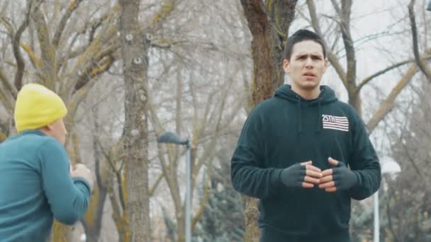 Entrenamiento de boxeo con entrenador al aire libre. Joven caucásico con capucha negra practicando golpes de sombra bajo la lluvia. El entrenador con gorra de punto amarillo enseña a esquivar con sentadillas . — Vídeo de stock