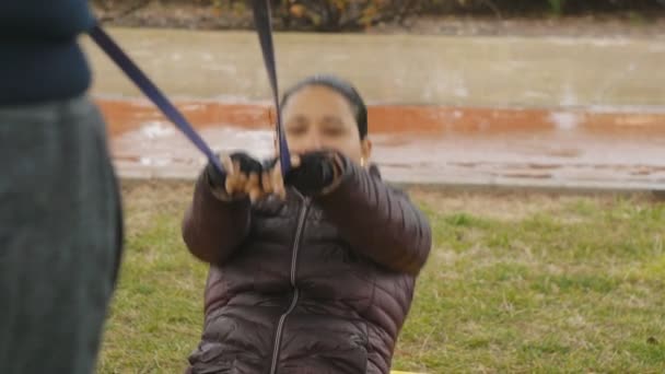 Un par de ejercicios al aire libre. Mujer hispana con chaqueta negra haciendo banda de resistencia se sienta con la ayuda del hombre en la lluvia de otoño como parte del programa de pérdida de peso . — Vídeo de stock