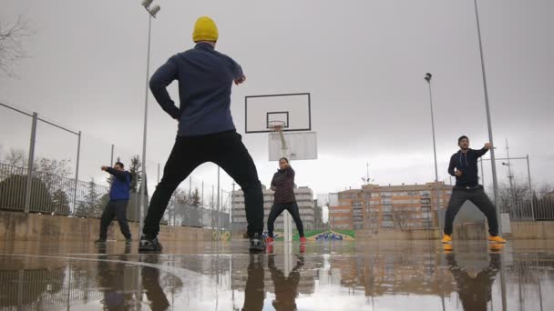 Street workout utbildning. Sport grupp unga multi etniska människor tränar tai chi stämplingar på utomhus basketplanen i regnet. Tränare med gul Stickad mössa förklarar korrekt rörelser. — Stockvideo
