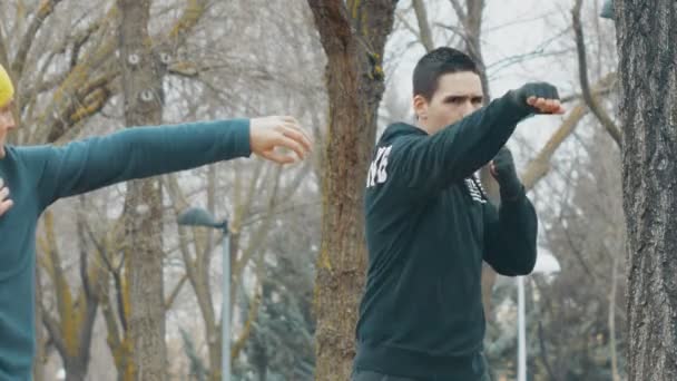 Entrenamiento de boxeo con entrenador al aire libre. Joven caucásico con capucha negra practicando golpes de sombra bajo la lluvia. El entrenador con gorra de punto amarillo enseña a perforar con sentadillas . — Vídeos de Stock