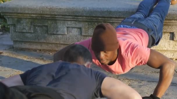 Treino com personal trainer ao ar livre. Homem de fitness preto e caucasiano fazendo flexões face a face com os pés elevados em um parque como parte de uma rotina de exercícios . — Vídeo de Stock