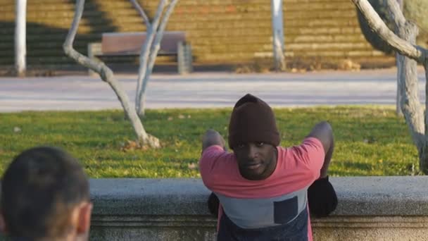 Träning med personlig tränare utomhus. Svarta och kaukasiska fitness man gör bänk dips ansikte mot ansikte med fötter som förhöjda i en park som en del av en workout rutin. — Stockvideo