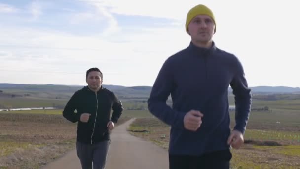 Entrenamiento con entrenador personal al aire libre. Dos hombres trotando en cámara lenta a lo largo de un camino rural sobre un fondo montañoso en otoño o primavera . — Vídeo de stock