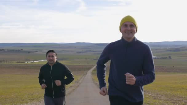 Entrenamiento con entrenador personal al aire libre. Dos hombres corriendo en cámara lenta sonriendo a lo largo de un camino rural sobre un fondo montañoso en otoño o primavera . — Vídeo de stock