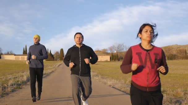 Workout mit Personal Trainer im Freien. zwei Männer und eine Frau laufen im Herbst oder Frühjahr in Zeitlupe die Landstraße hinunter. — Stockvideo