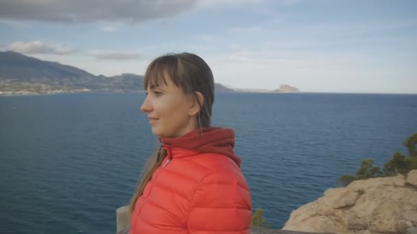 Młoda kobieta na tle morza. Uśmiechający się kaukaski kobieta Brunet w czerwona kurtka Bluza z kapturem i bańki czekamy od góry. — Wideo stockowe