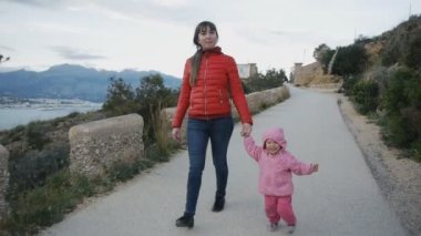 Şirin beyaz bebek sahil manzara arka plan üzerinde kamera takip etmeye anneler elini tutmadan çalışır gülüyor başlıklı. Mutlu çocukluk kavramı.