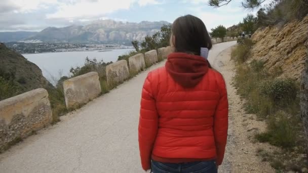 Вид ззаду на дівчину в червоній куртці, що йде по гірській дорозі на морському і гірському пейзажному фоні . — стокове відео