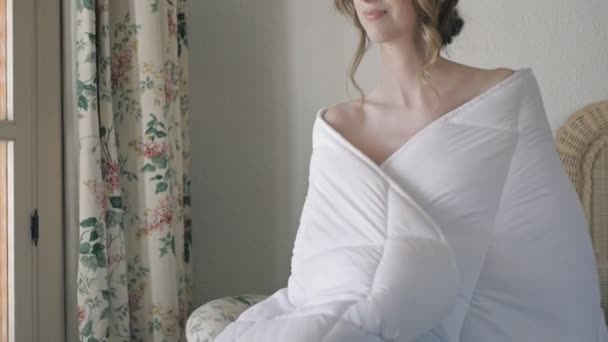 A fehér takaróba csomagolt fiatal nő otthon ül egy székben.. — Stock videók
