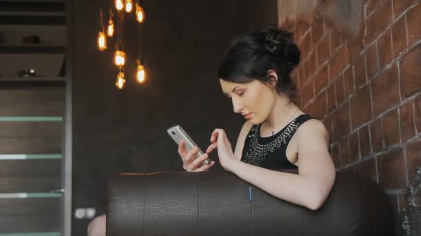 Μελαχρινή κοπέλα απολαμβάνει ένα τηλέφωνο σε ένα δερμάτινο καναπέ — Αρχείο Βίντεο