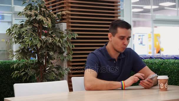 Un joven con un simbolismo LGBT navegando por un teléfono móvil en un café — Vídeos de Stock