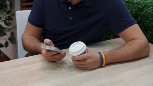 Un jeune homme avec un symbole arc-en-ciel dans un café, utilise un téléphone — Video