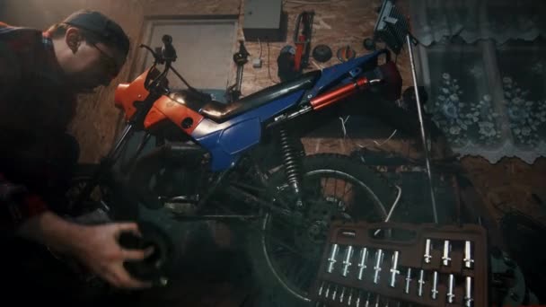 Механик в красной рубашке ремонтирует двигатель мотоцикла в гаражной мастерской — стоковое видео