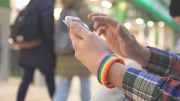 Un hombre con un símbolo LGBT que lleva una pulsera usa un teléfono en un centro comercial Fotos De Stock Sin Royalties Gratis