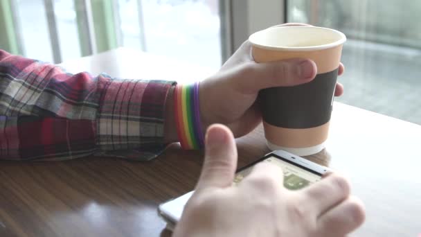 Mens mãos com símbolo pulseiras LGBT, em um café, uma xícara de café e um telefone celular — Vídeo de Stock