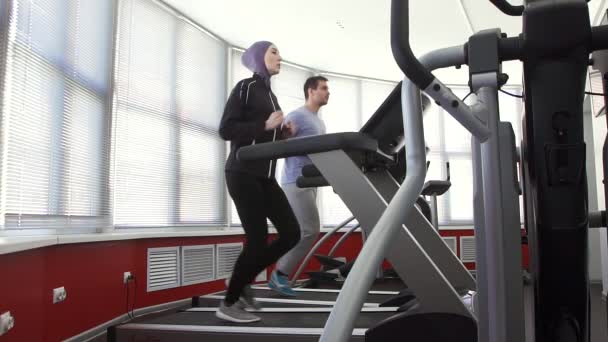 Sportlerin im Hidschab im Laufsimulator und ein Mann im Fitnessstudio — Stockvideo