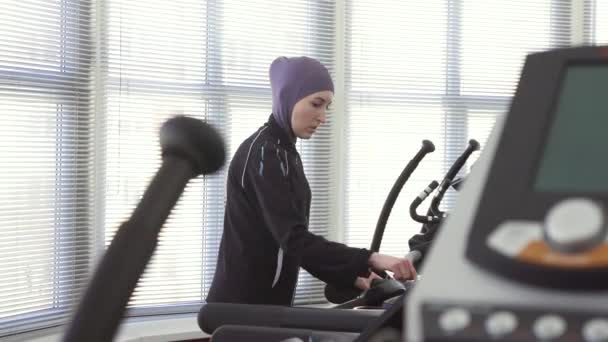 Ragazza in hijab, riscaldamento ambulante su un simulatore di corsa in palestra, allenamento di fitness — Video Stock