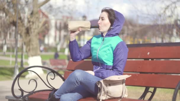 Vrouw drinkt uit een fles in een papieren zak in een park op een bankje — Stockvideo