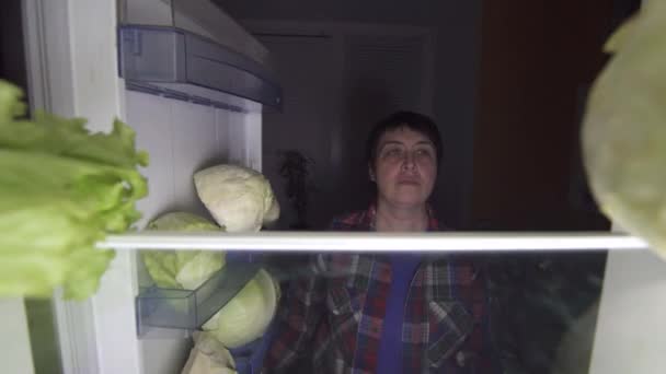 Mujer de 40 años en una dieta abre el refrigerador por la noche y mira una ensalada verde — Vídeos de Stock