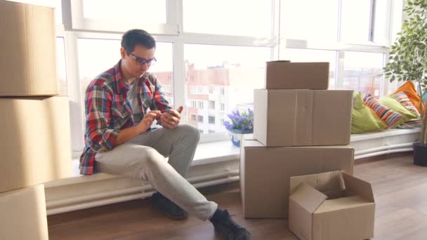 Hombre joven utilizado en el teléfono sentado en el nuevo apartamento de mudanza en — Vídeos de Stock